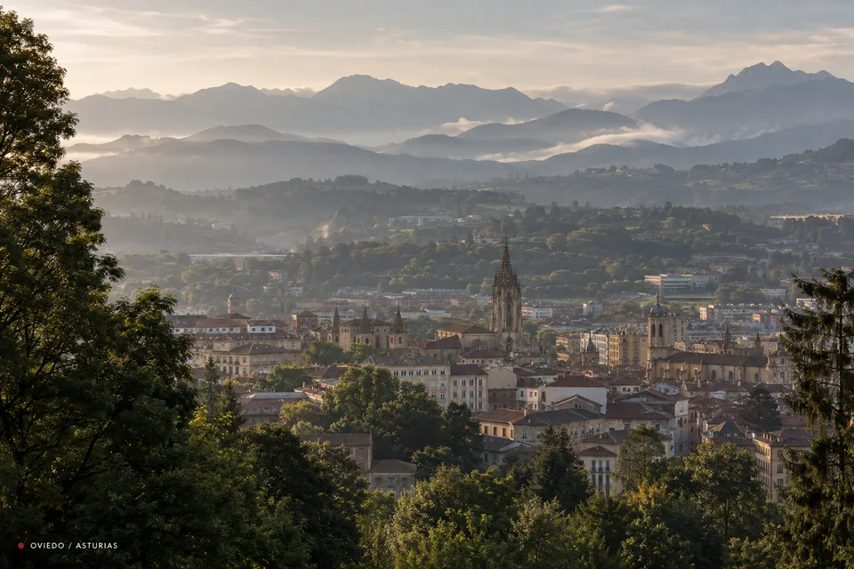 Editorial view of Oviedo among trees and the mountains of Asturias, with a discreet location marker.
